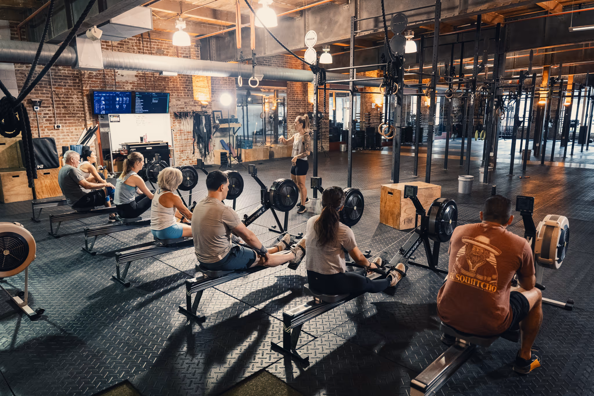 Group of adults exercising on rowing machines in a gym with an instructor standing and guiding them.