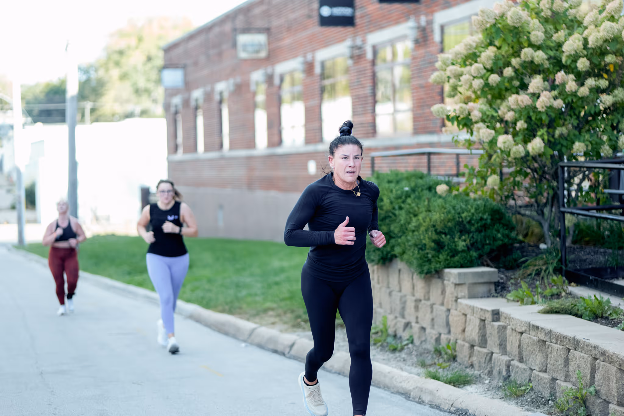 Three women running outdoors on a street beside a brick building with greenery.