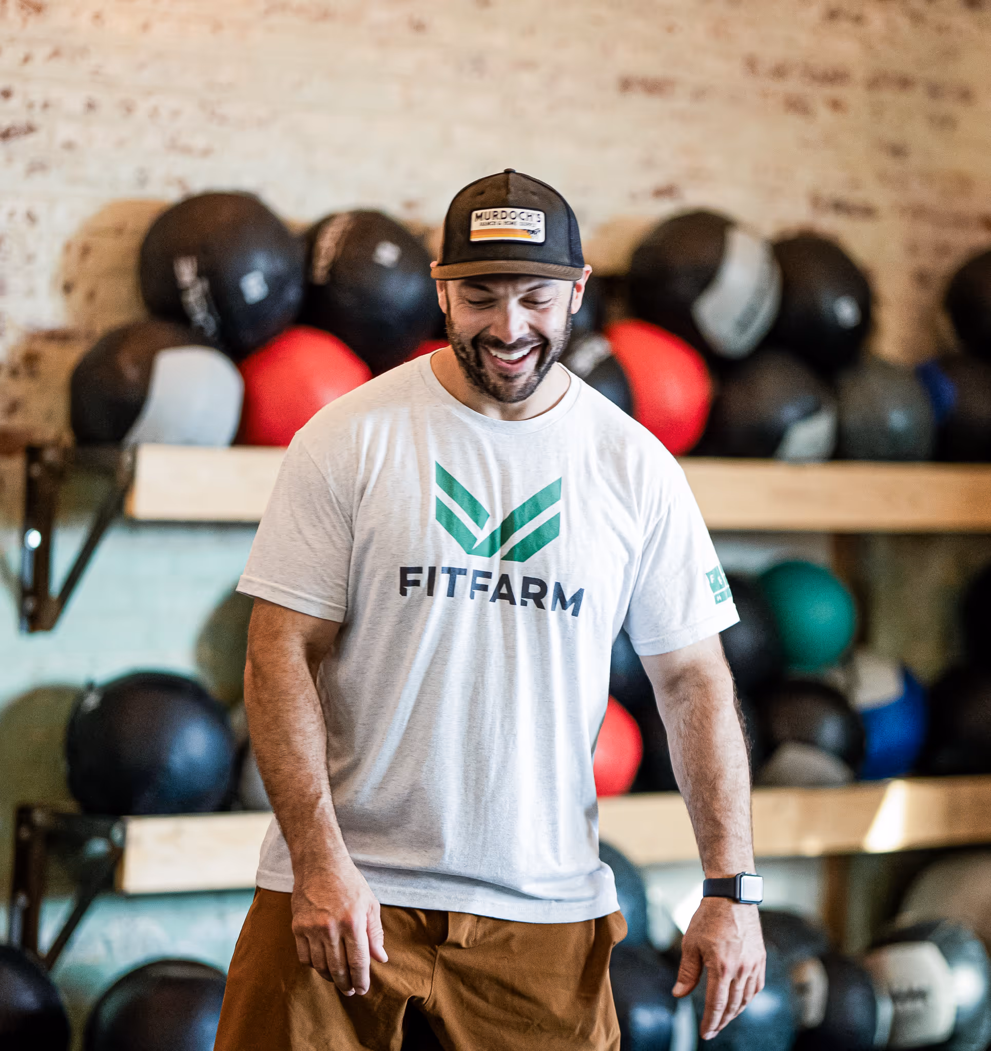 Smiling man wearing a FITFARM t-shirt and cap standing in front of shelves with medicine balls in a gym.