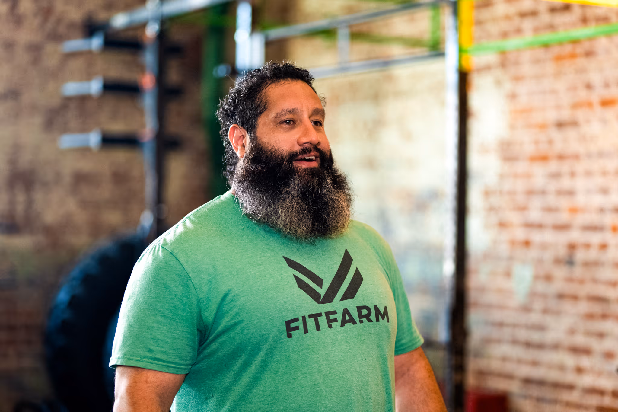Bearded man wearing a green FitFarm t-shirt standing in a gym with blurred equipment and brick wall background.