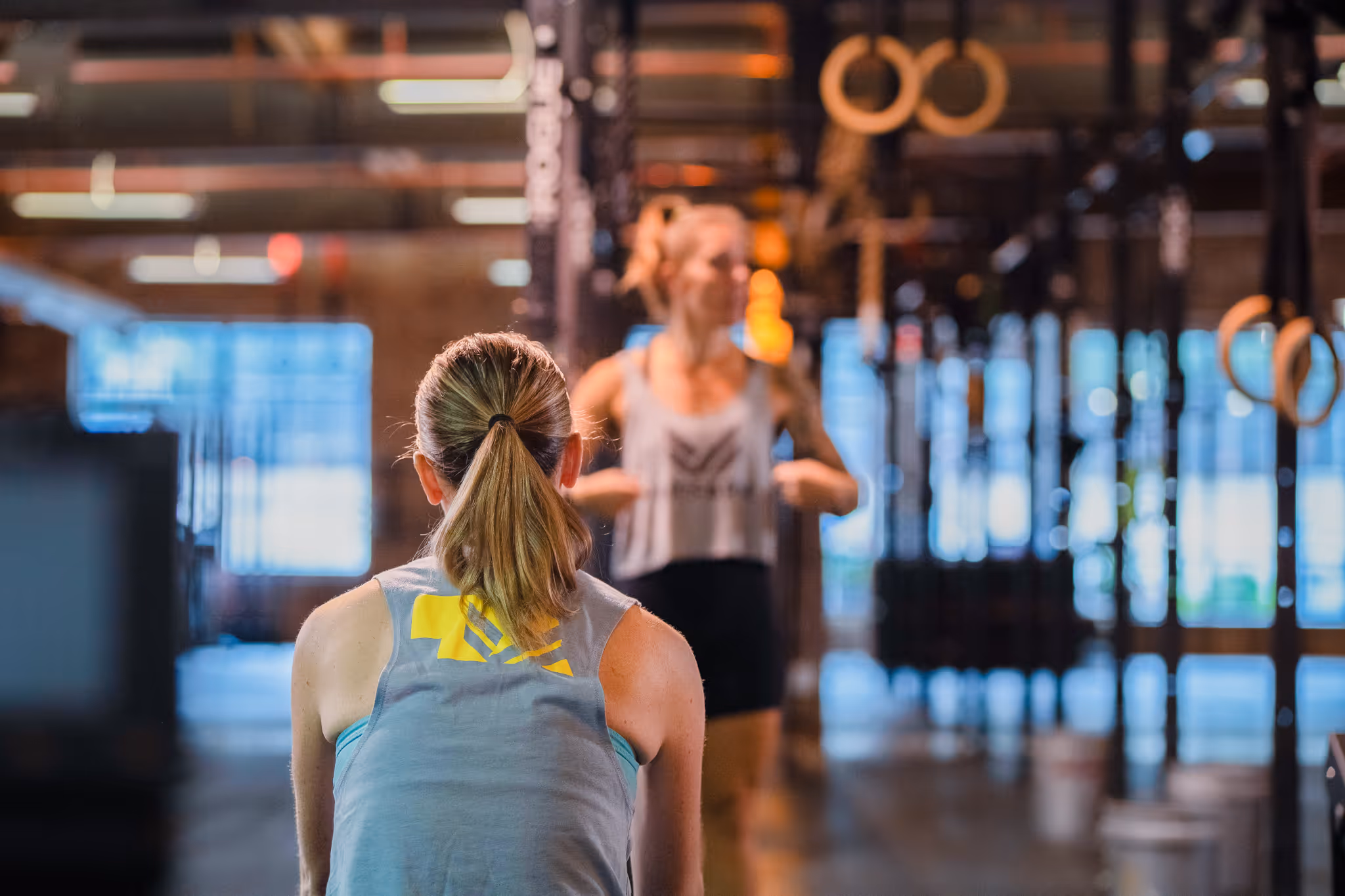 Two women in a gym with one facing away and the other standing in front with arms bent.