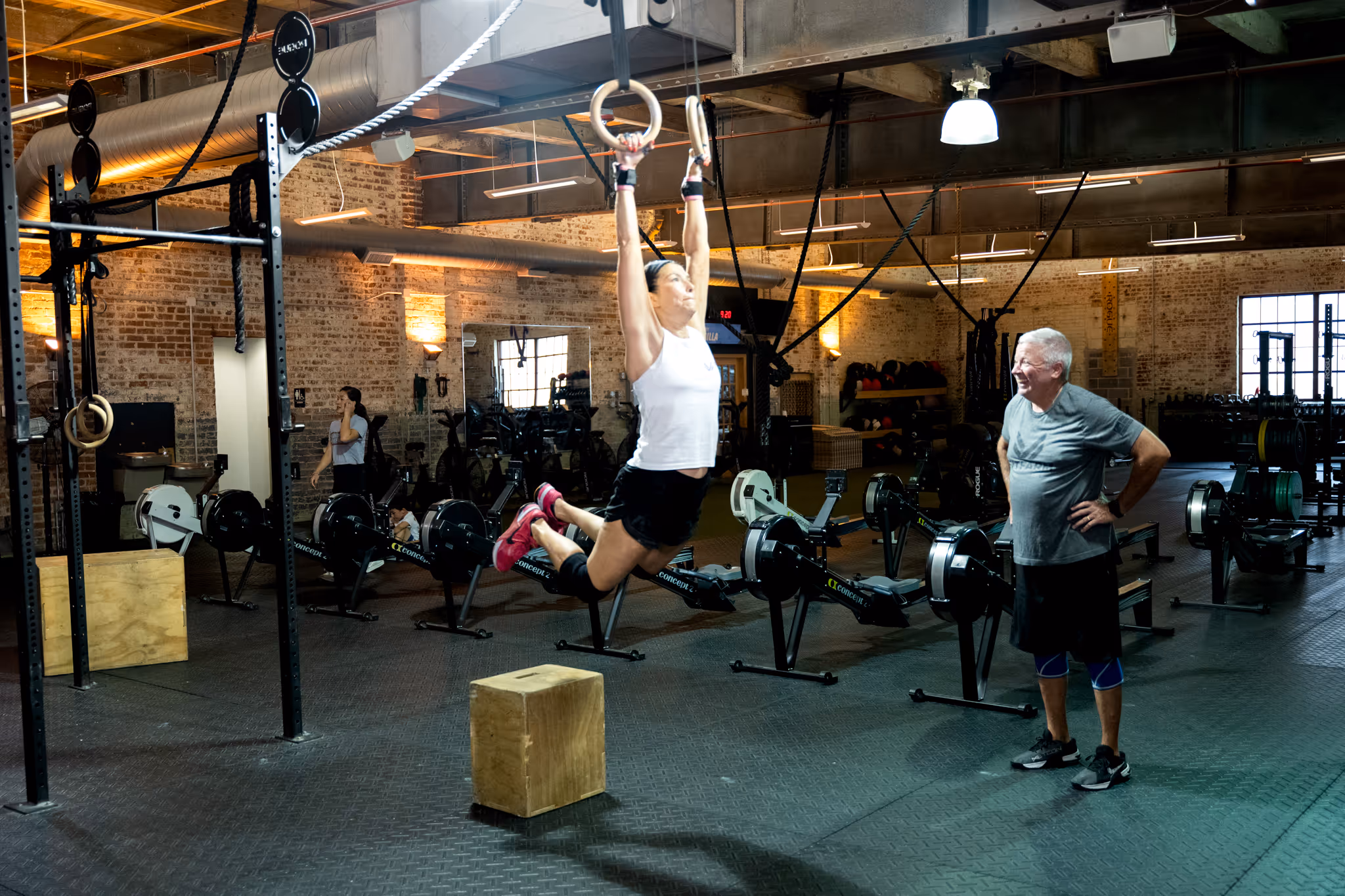 Woman in white tank top and black shorts performing a ring muscle-up in a gym while an older man watches and smiles.