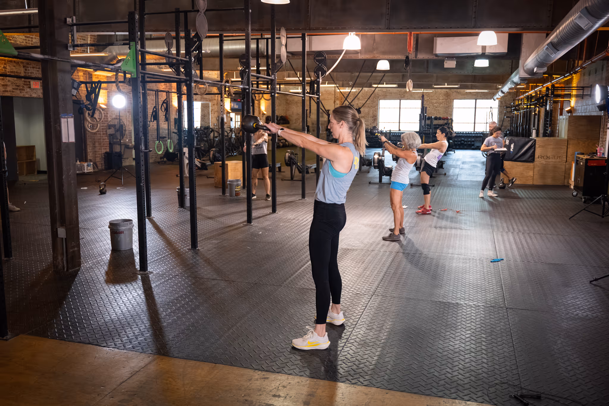 Group of women exercising with kettlebells in a spacious gym with industrial decor.