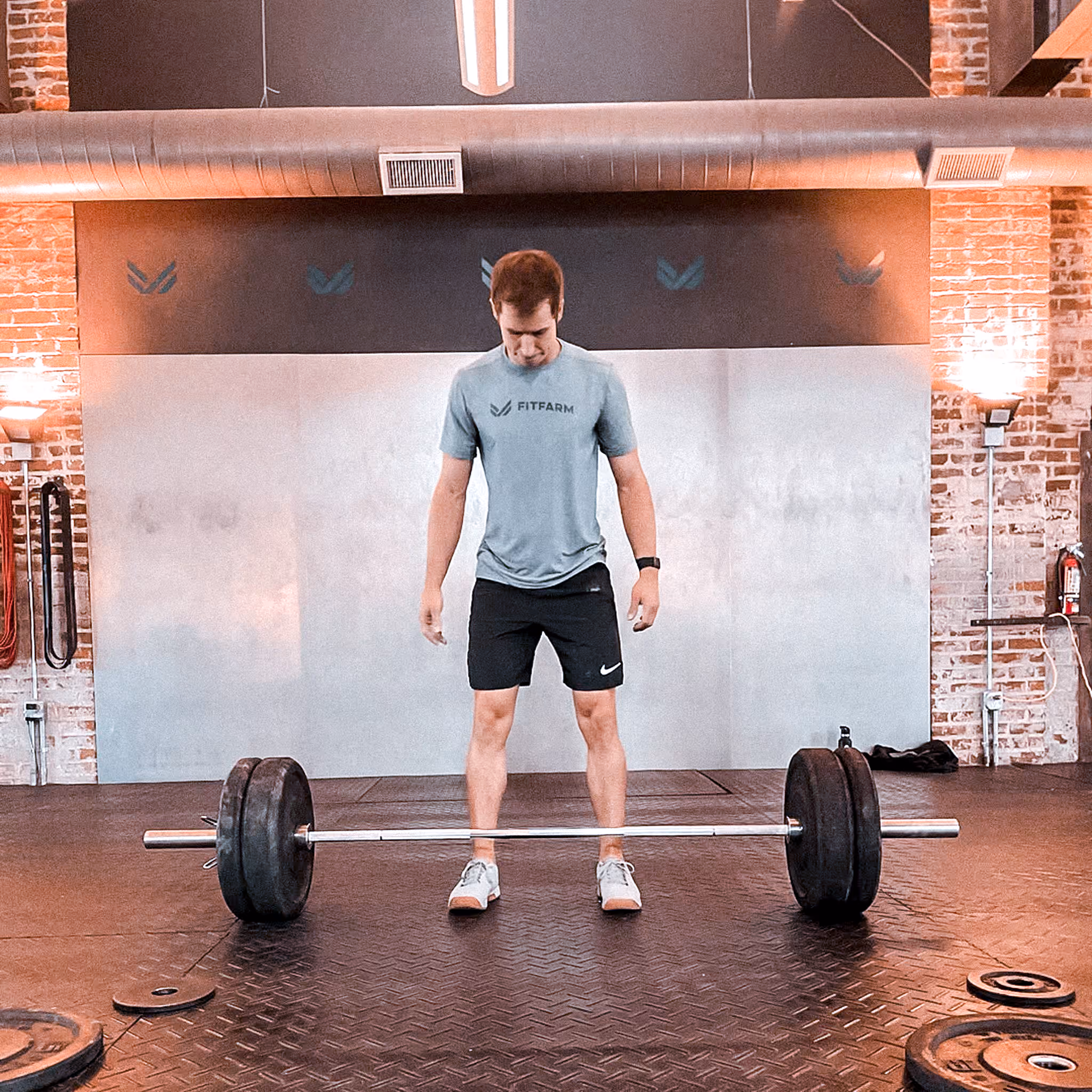 Man in workout attire standing before a loaded barbell in a gym with brick walls and rubber flooring.
