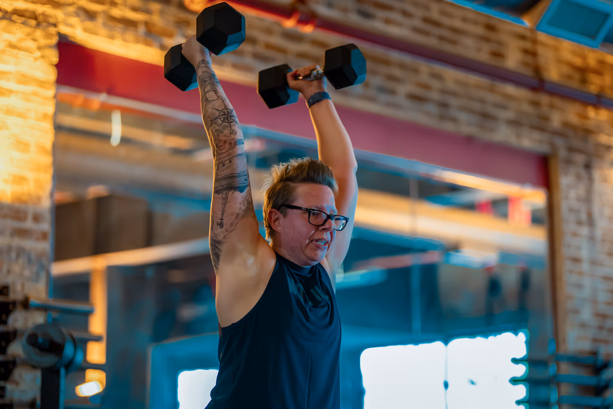 Person wearing glasses lifting black dumbbells overhead in a gym with brick walls and large mirror.