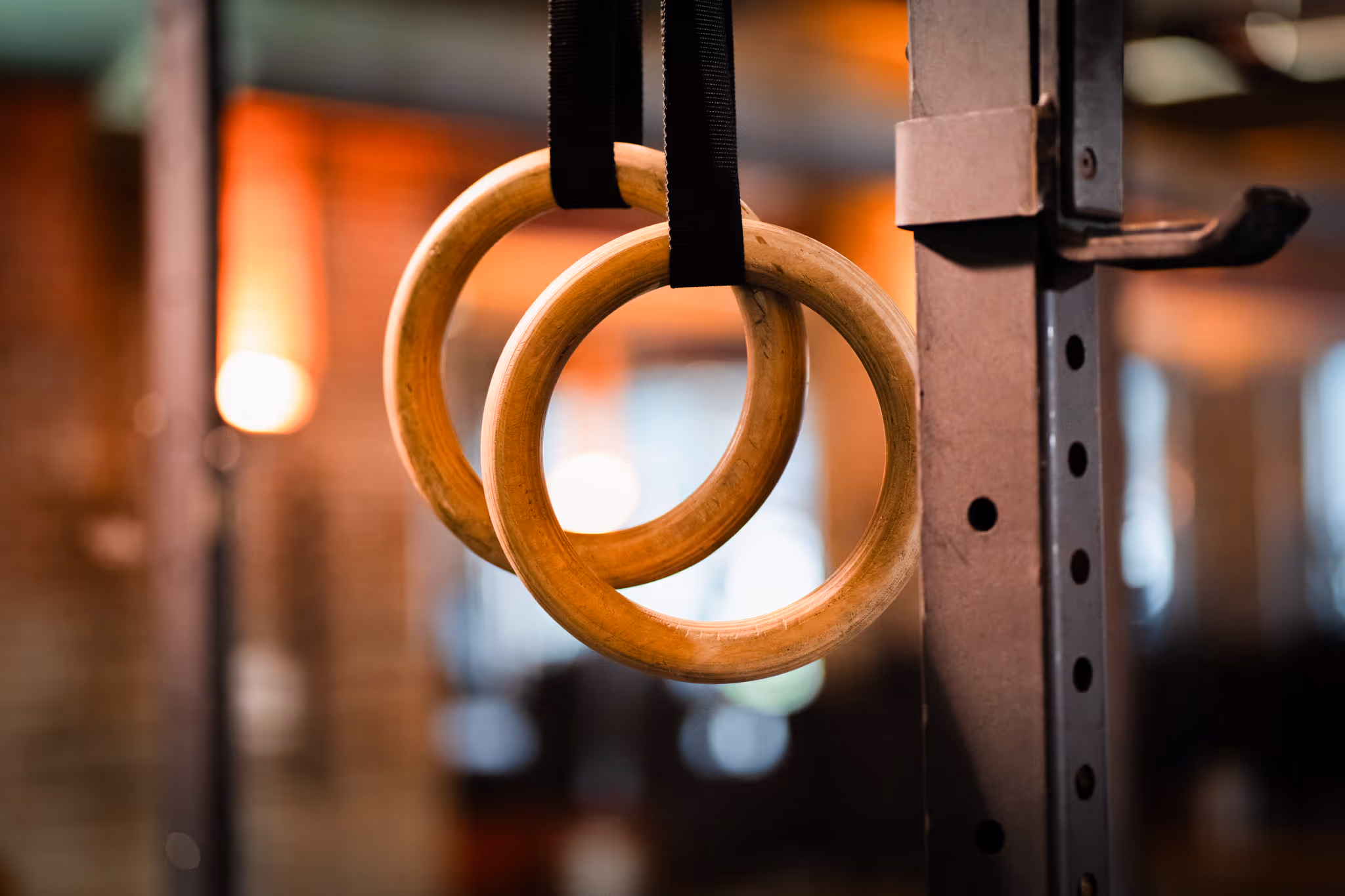 Close-up of wooden gymnastic rings hanging from black straps attached to a metal frame in a gym.