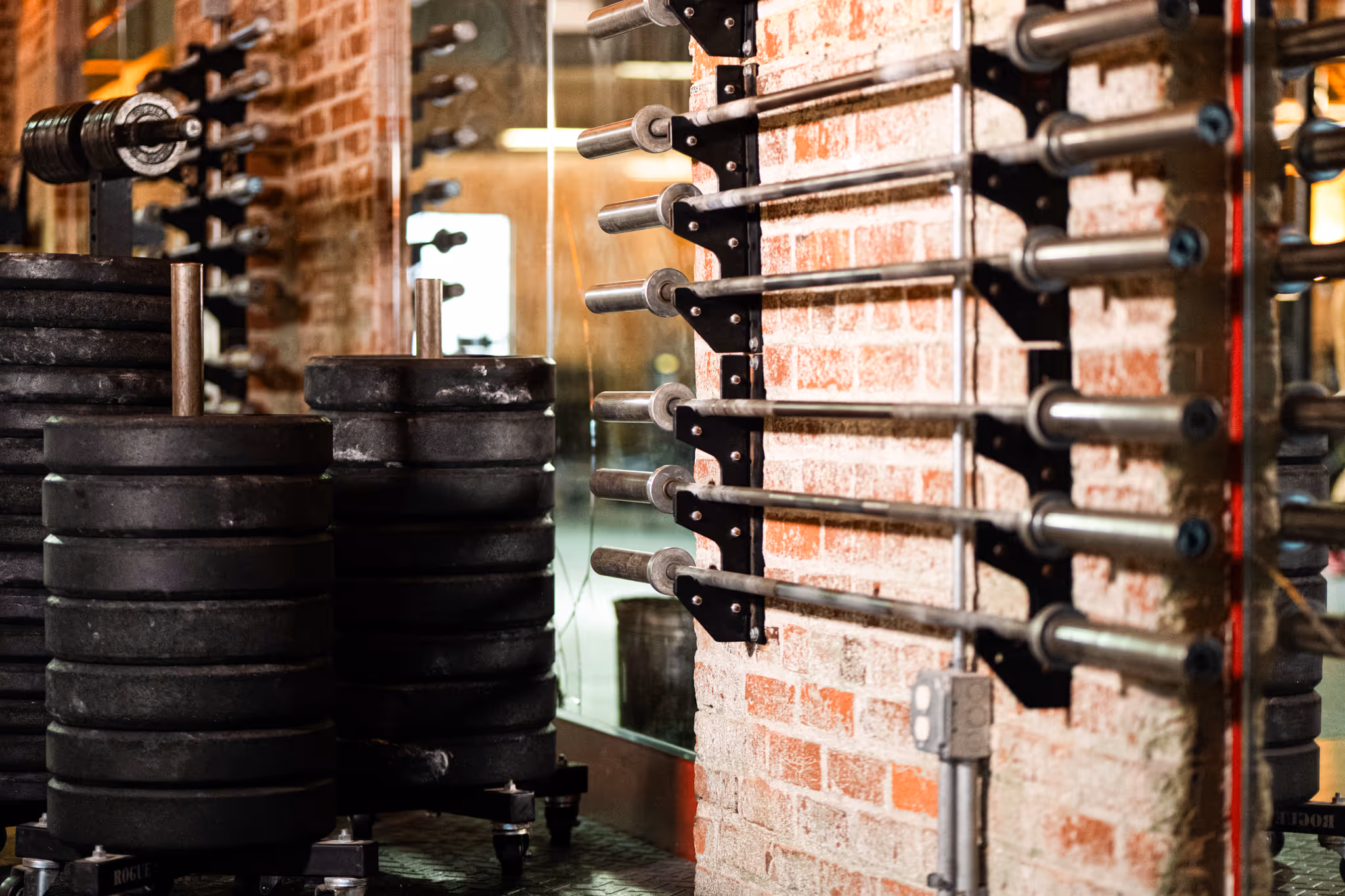 Rack with multiple barbell bars mounted on a brick wall in a gym beside stacks of weight plates.