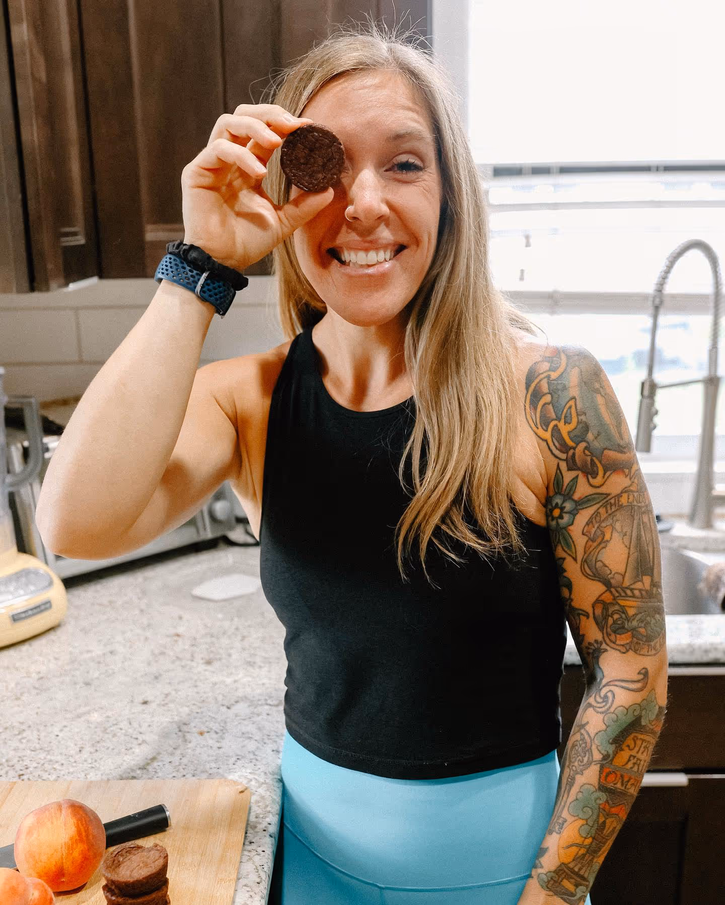Smiling woman with tattoos in kitchen holding a cookie over one eye.