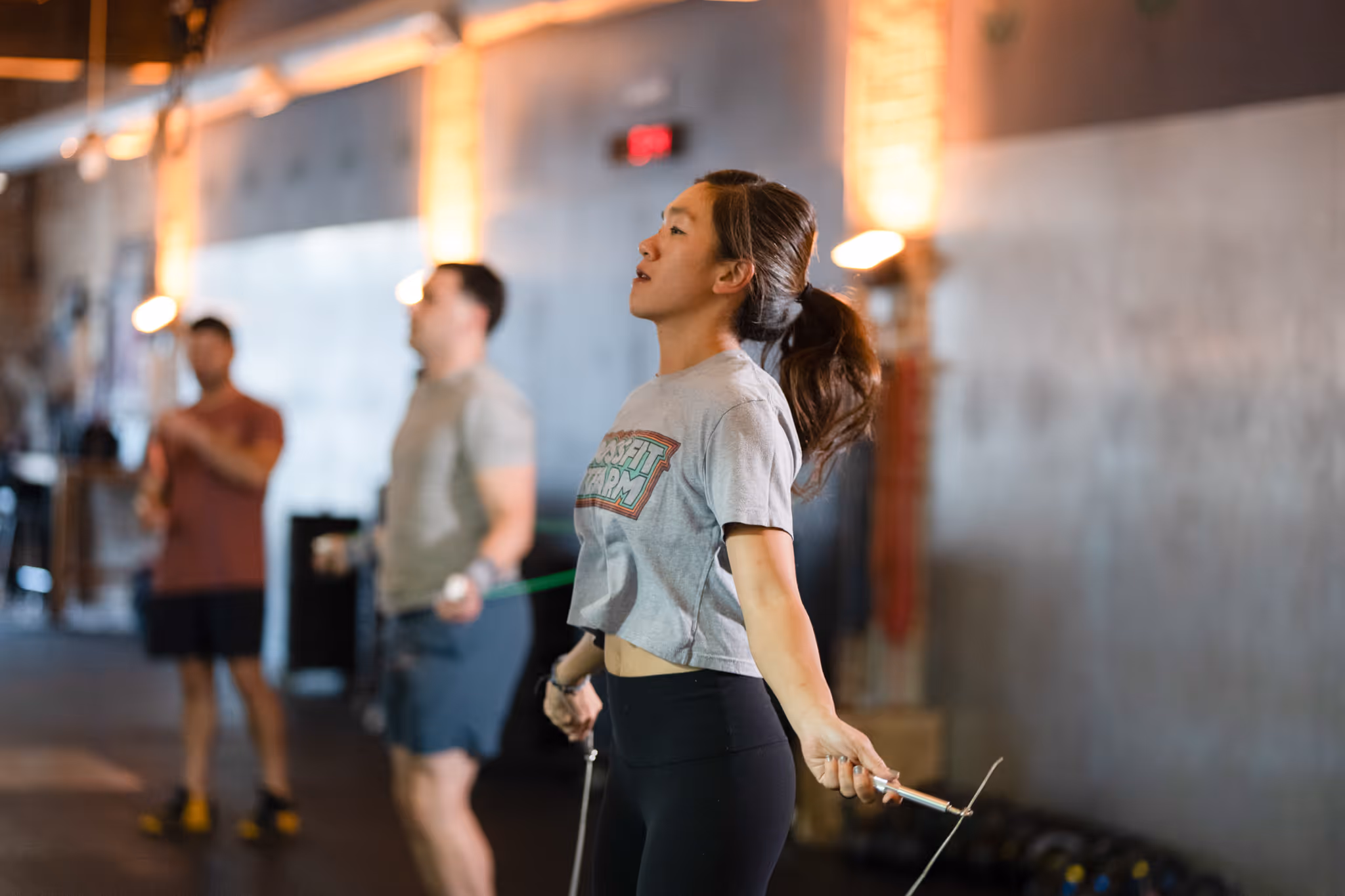 Three people exercising with jump ropes in a gym, focusing on a woman in a gray t-shirt and black leggings.