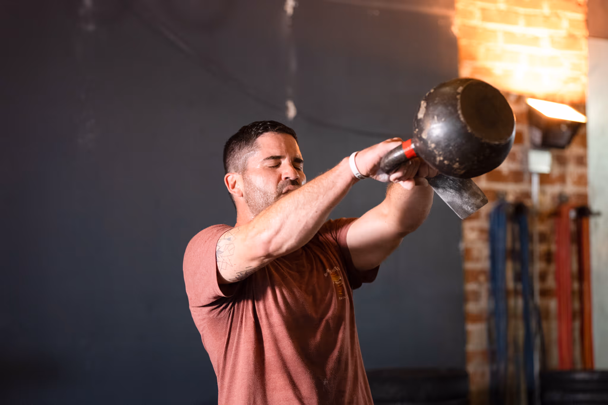 Man in a brown t-shirt lifting a kettlebell during a workout in a gym.