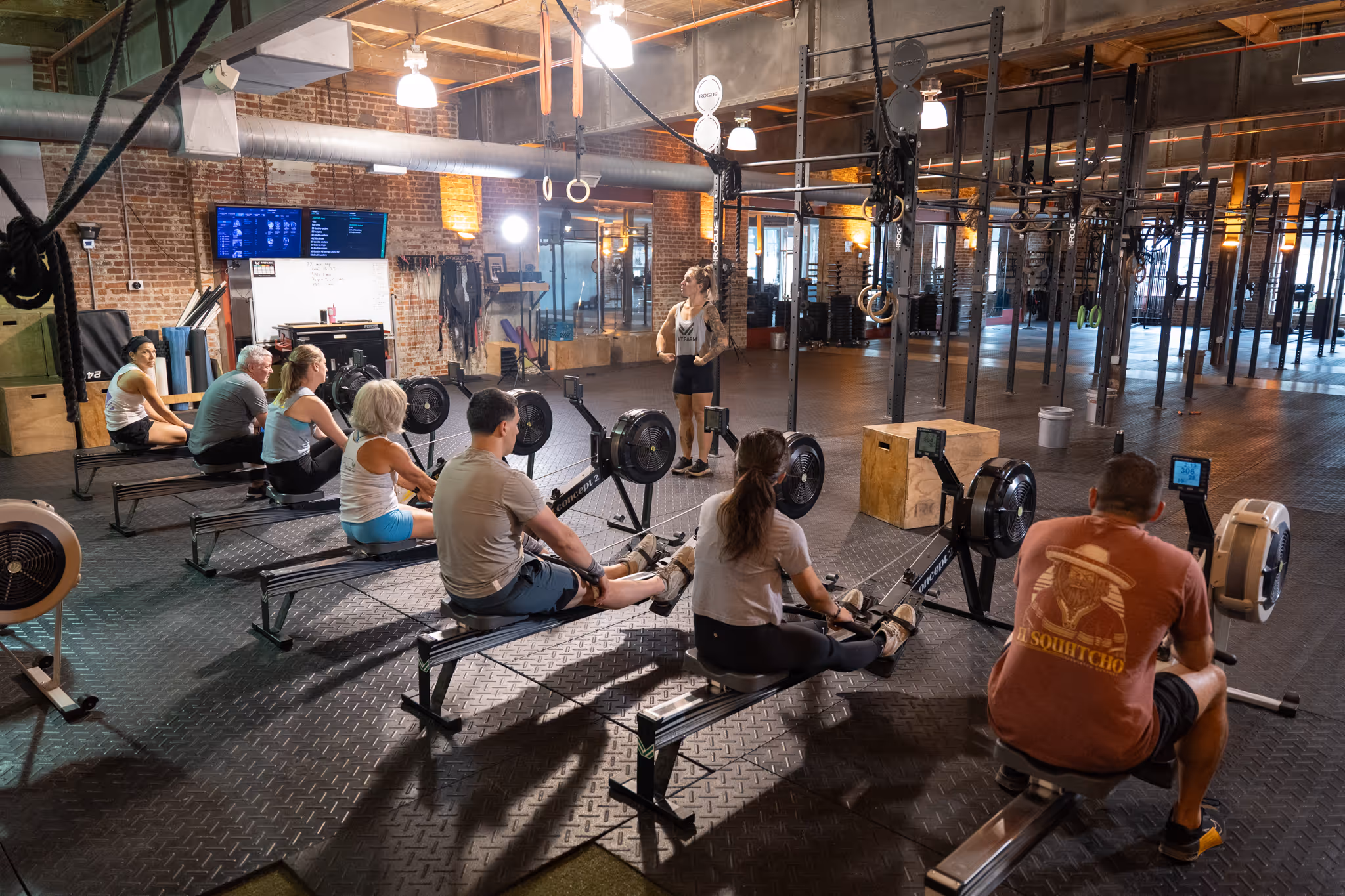 Group fitness class with seven people using rowing machines in an industrial-style gym.