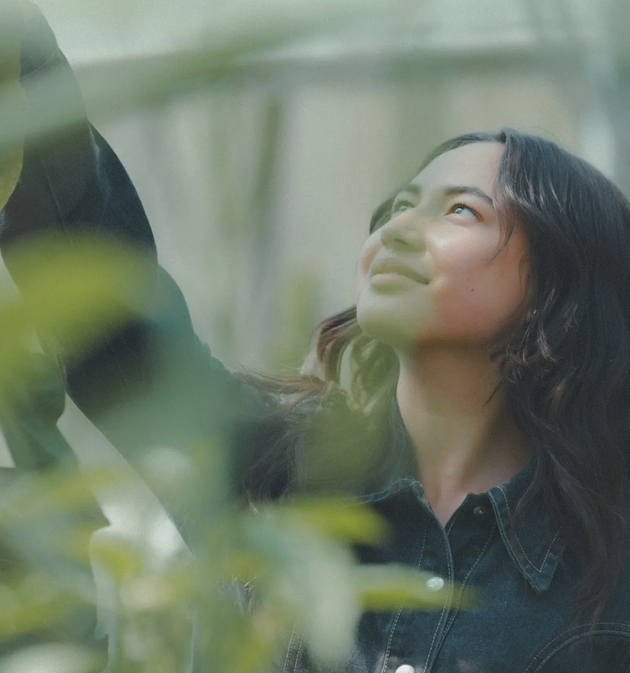 A young woman with long dark hair, wearing a denim shirt, stands outdoors among green plants, looking up and reaching her arm upward with a calm expression—embracing the benefits of science-based care and Glutathione for a healthier lifestyle.