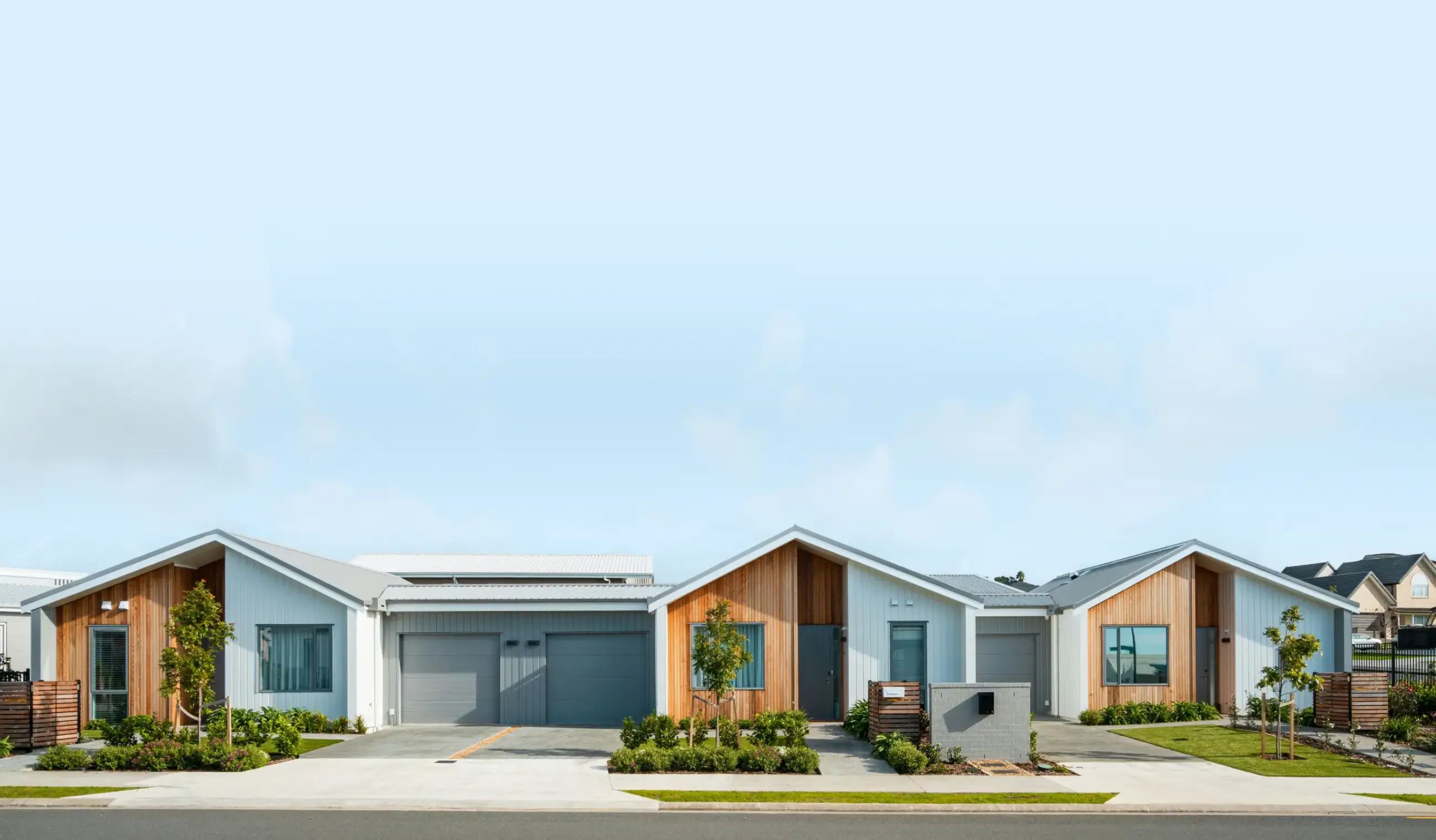 Row of modern single-family homes with wood paneling, grey roofs, garages, and landscaped front yards under a clear blue sky.