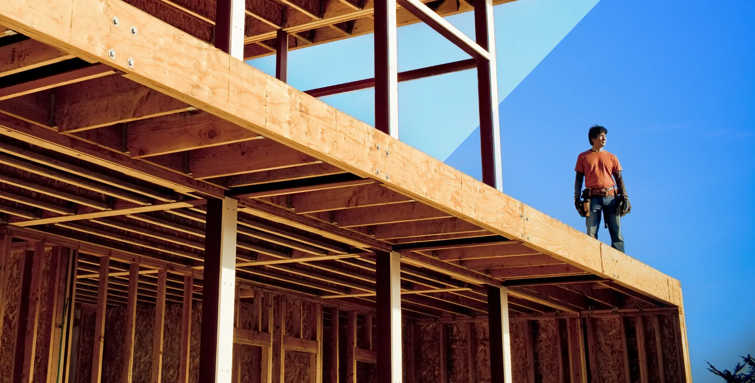 Man wearing tool belt standing on the wooden frame of a house under construction against a blue sky.