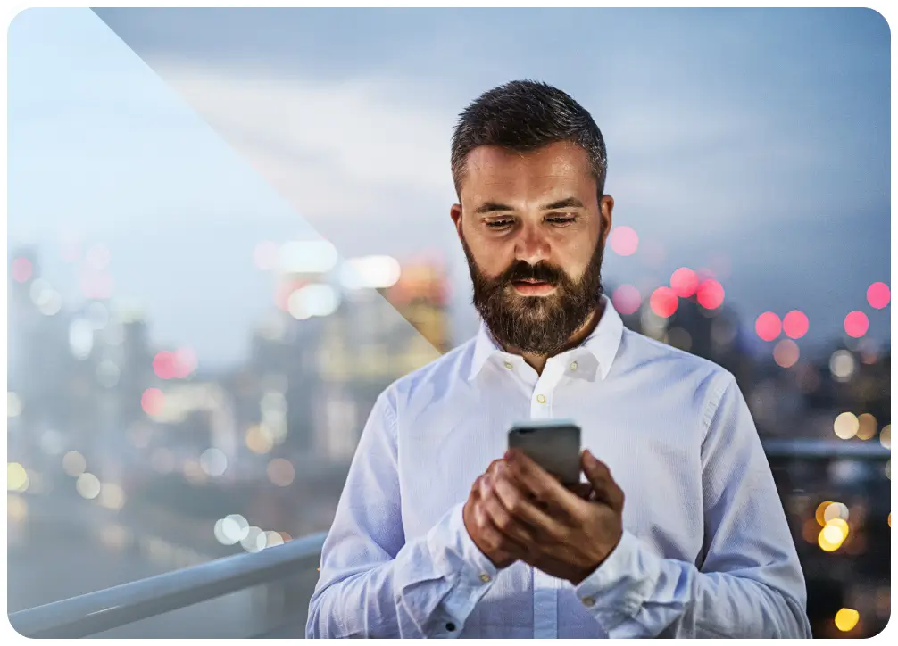 Bearded man in a white shirt looking at his smartphone with a city skyline blurred in the background during dusk.