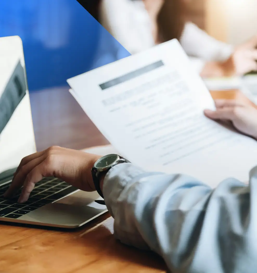 Person reviewing a document while typing on a laptop keyboard.