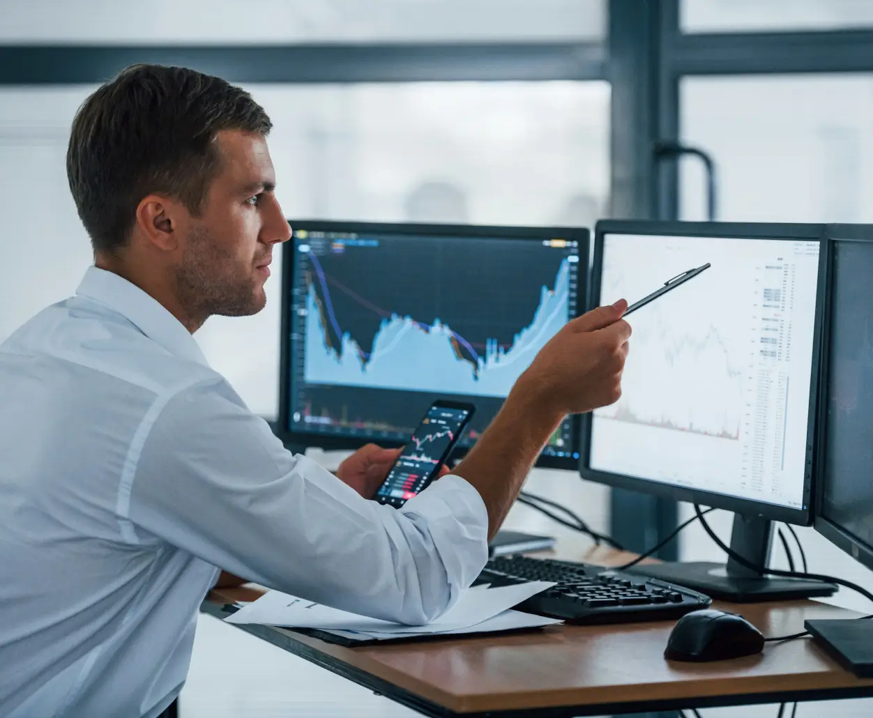 Man in a white shirt analyzing financial charts on dual computer monitors while holding a smartphone and pointing at one screen with a pen.