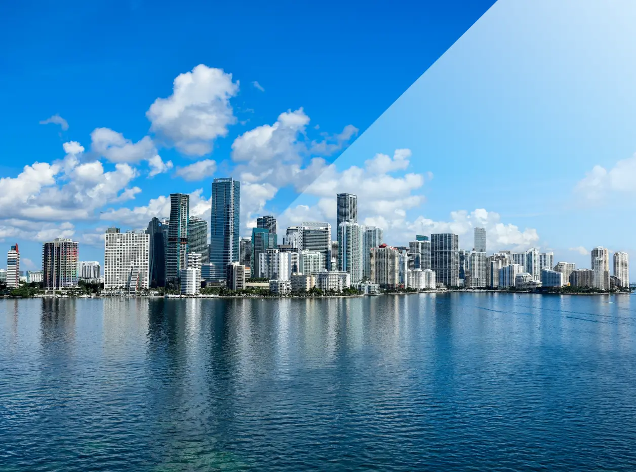 Skyline of a city with tall buildings reflected in calm water under a blue sky with scattered clouds.