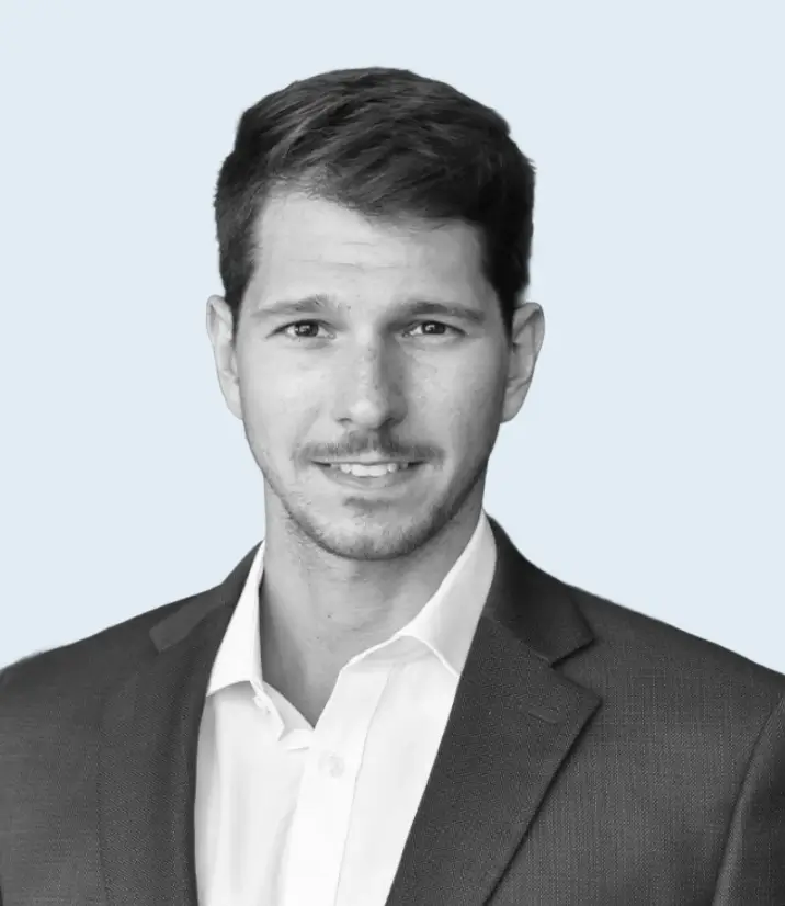 Portrait of a young man with short brown hair wearing a dark suit jacket and white shirt against a light blue background.
