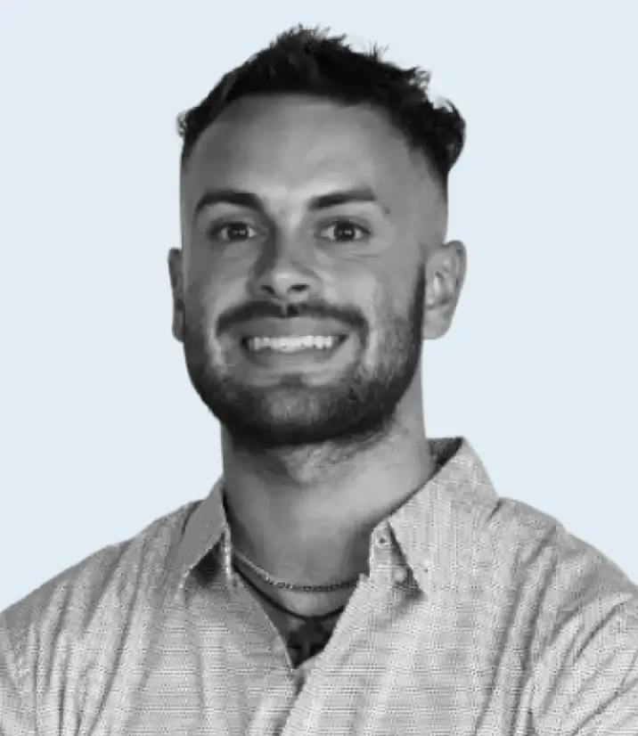 Smiling man with short curly hair and beard wearing a checkered shirt and a necklace against a light background.