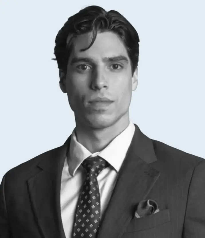 Portrait of a serious young man wearing a dark suit, patterned tie, and white dress shirt against a light background.