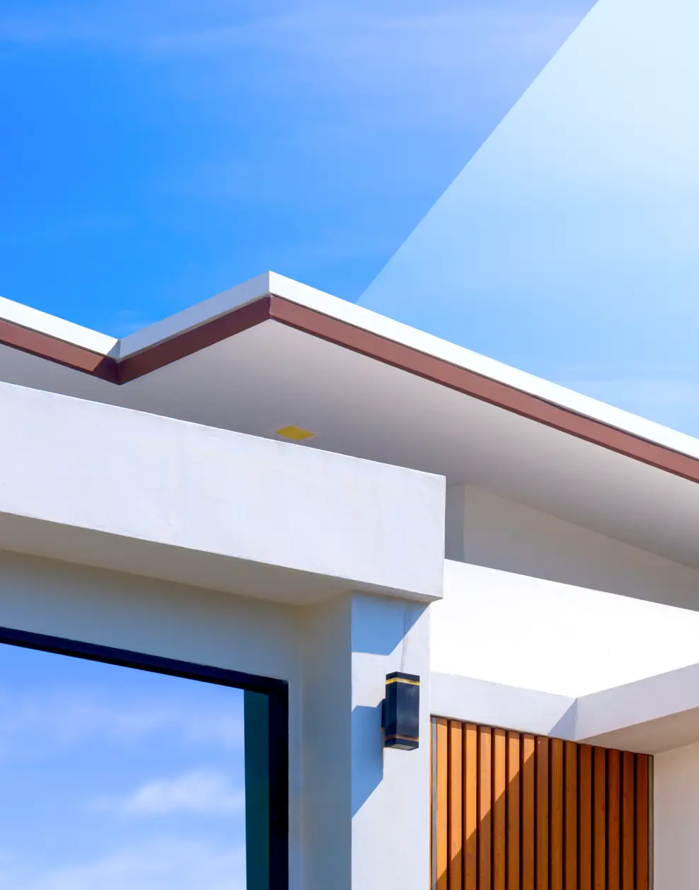 Modern luxury home exterior with white walls, wooden panel accents, and a black outdoor light fixture under a clear blue sky.