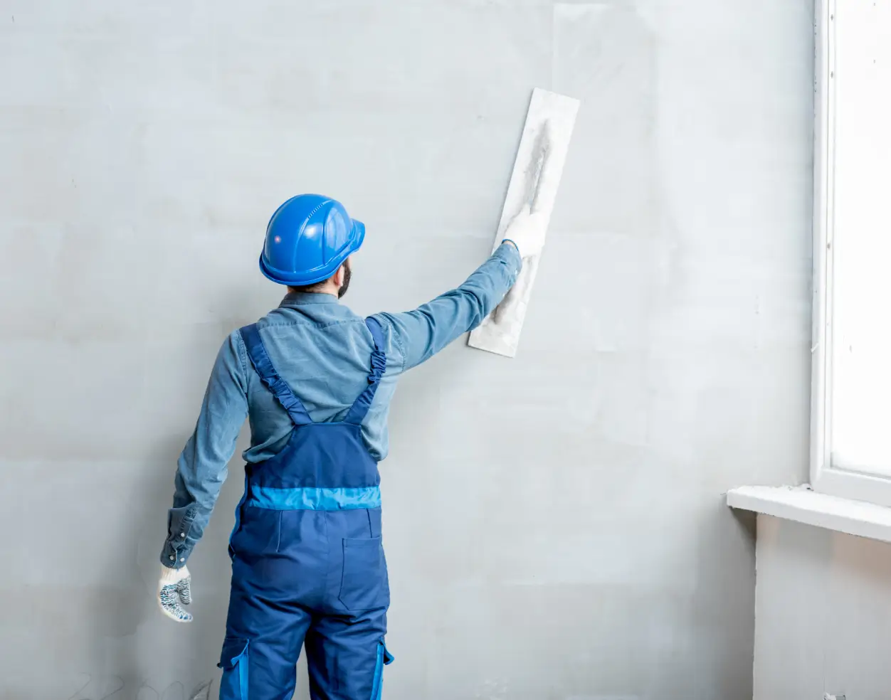 Construction worker wearing blue overalls and hard hat smoothing a wall plaster near a window.