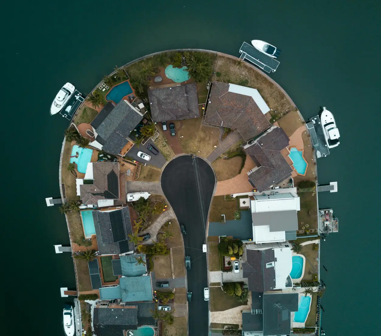 Aerial view of a circular cul-de-sac surrounded by waterfront houses with swimming pools and boats docked along the water edges.
