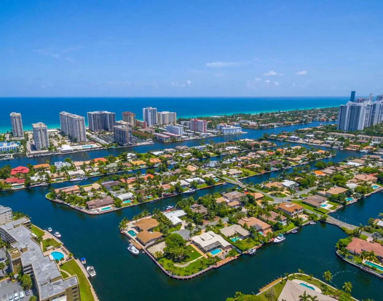 Aerial view of a coastal city with waterways, residential houses with pools, high-rise buildings, and a clear blue ocean under a sunny sky.
