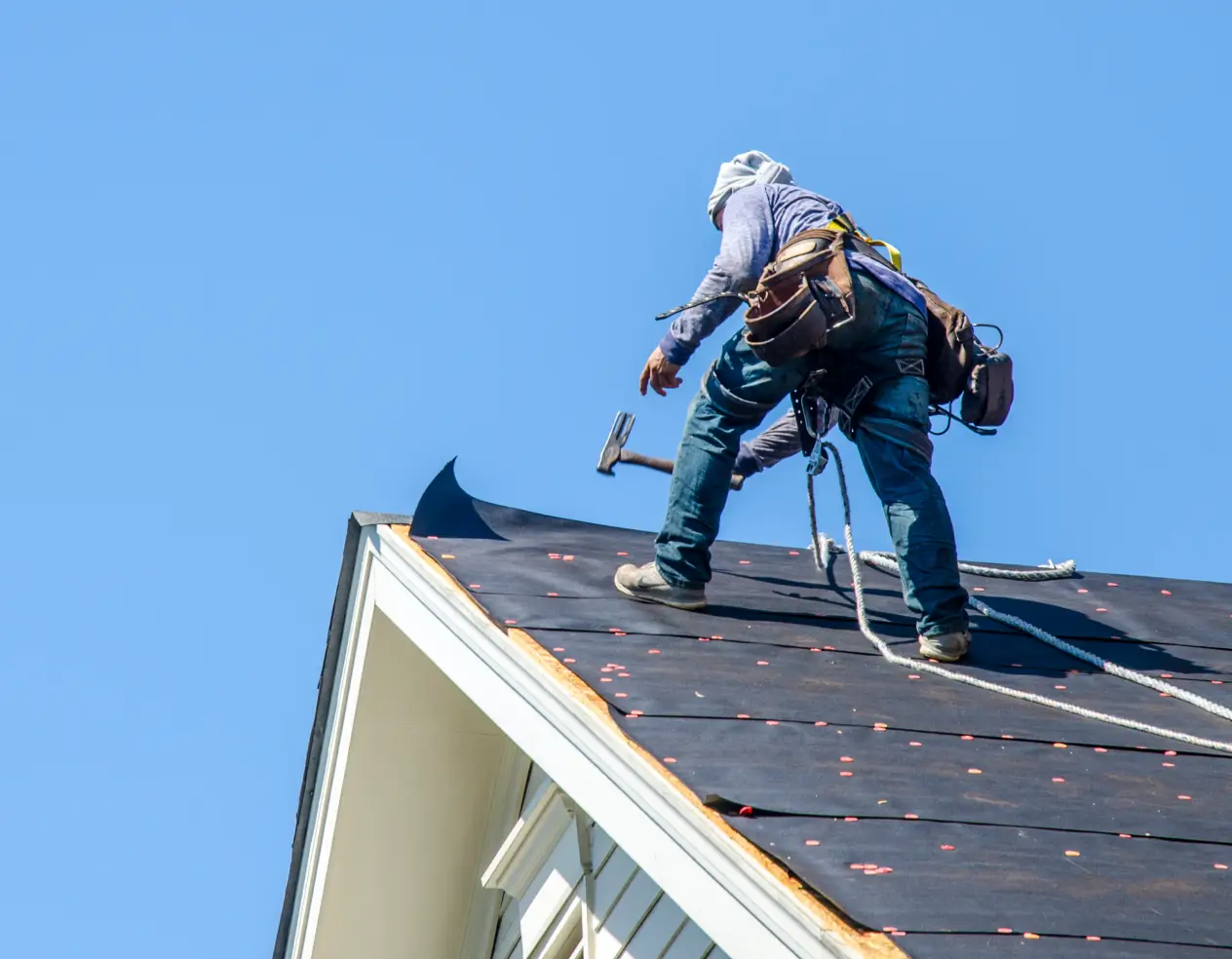 Roofer wearing safety gear and tool belt using a hammer on a steep roof under clear blue sky.