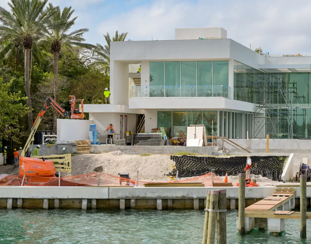 Modern luxury home under construction by the waterfront with construction workers, scaffolding, and equipment.