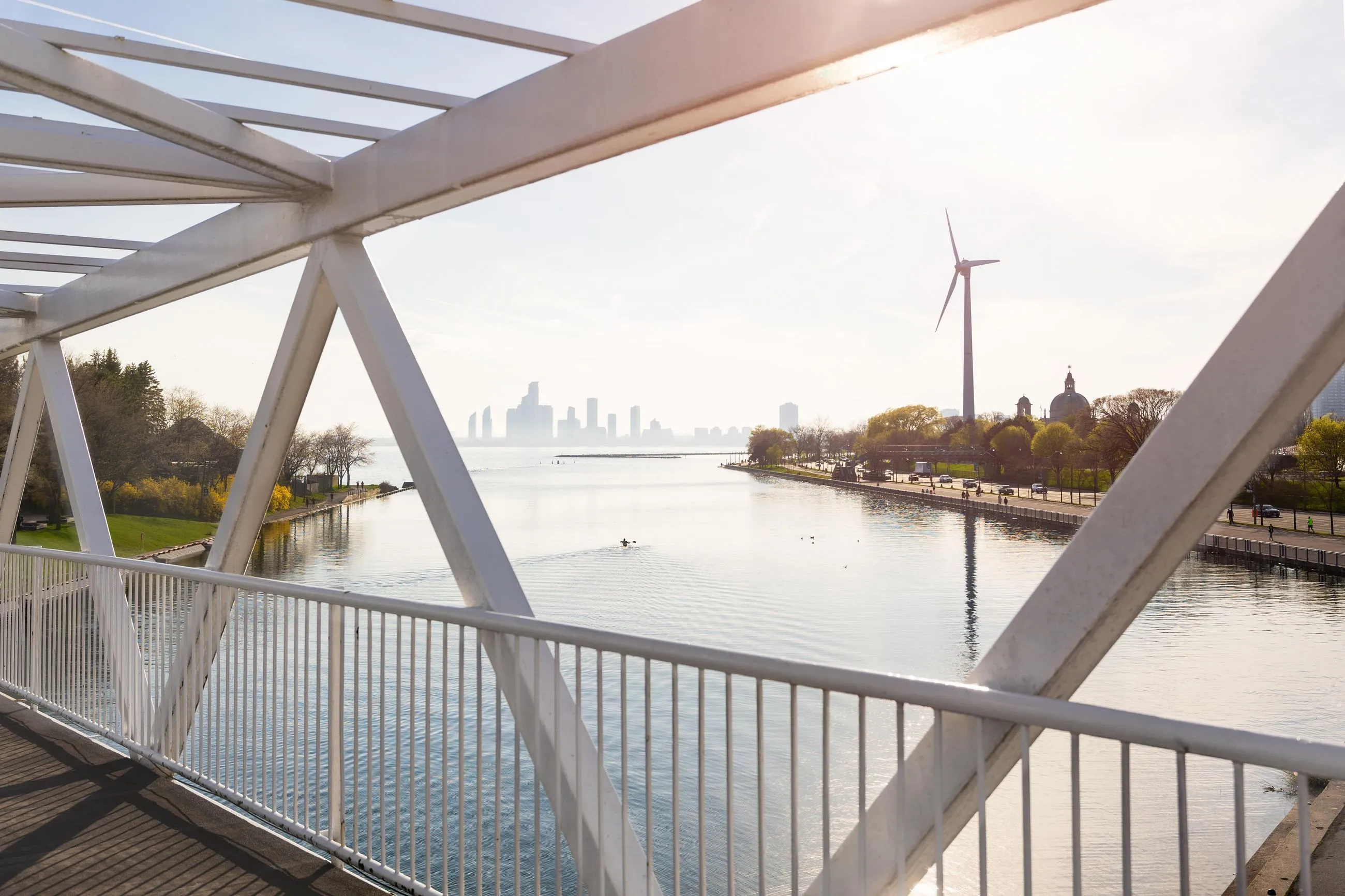 A photo of a water front capturing a park and a city's skyline taken from in between the metal frame of a bridge.