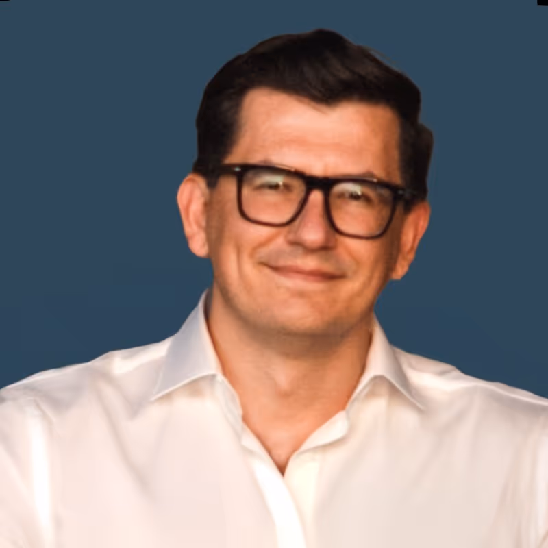 
A headshot of a man with dark, neatly combed hair and black-framed glasses. He is wearing a white collared shirt and is smiling faintly, looking directly at the camera. The background is a solid, dark blue color.