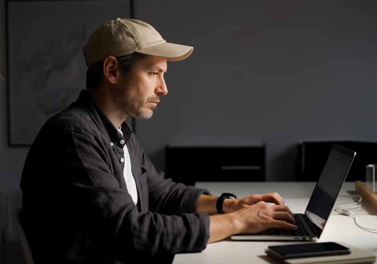 A man with short hair and a light-colored baseball cap is seated at a white desk, intently typing on a laptop. He is wearing a dark, possibly charcoal, button-up shirt over a white t-shirt. On his left wrist, he has a dark watch. The background is a plain, dark gray wall with a framed picture visible on the right. The lighting suggests a focused, indoor environment, with light illuminating his face and the laptop. A smartphone and another object are visible on the desk to his right.