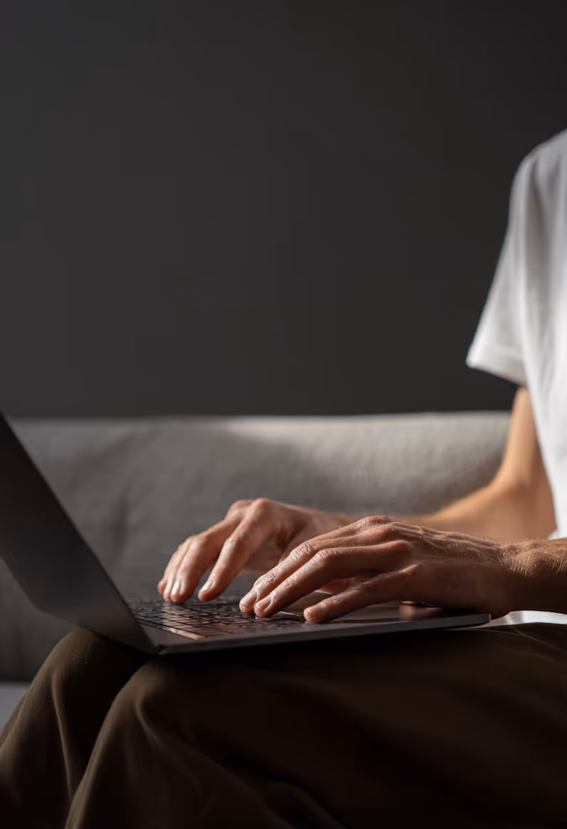 A close-up, cropped shot shows a person's hands typing on a laptop. The person is wearing a white t-shirt and dark-colored pants, and is seated on what appears to be a light gray couch. The background is a dark gray wall, and there's a strong, directional light source (likely sunlight) casting shadows and highlighting the hands and parts of the laptop.