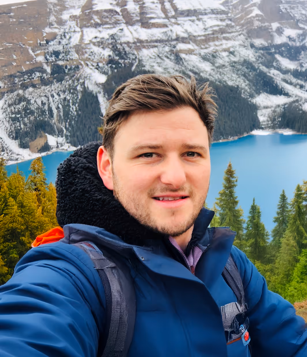 A man with light brown hair and a beard is taking a selfie outdoors, smiling at the camera. He is wearing a blue winter jacket with a black, possibly furry, collar and a backpack. Behind him is a stunning landscape featuring snow-capped mountains, green evergreen trees, and a vibrant turquoise or blue lake.