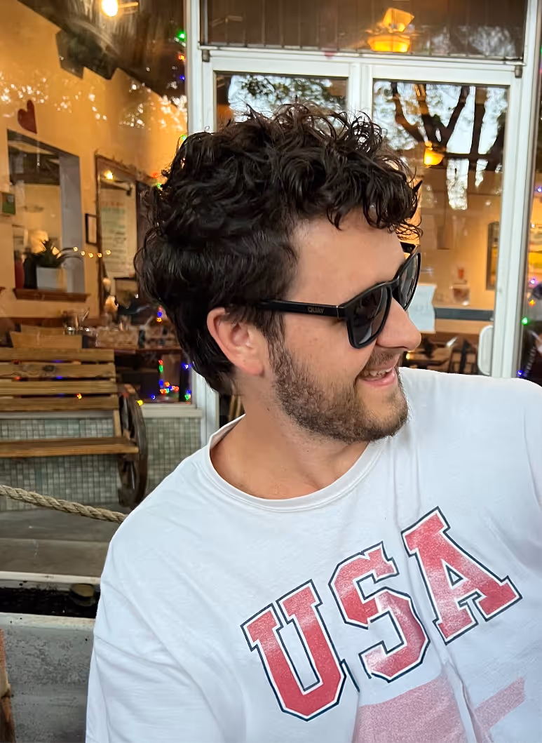 A man with dark, curly hair and a beard is smiling and looking to his right (out of frame). He is wearing dark sunglasses and a white t-shirt with "USA" printed in red and blue letters across the front. He appears to be seated outdoors at a cafe or restaurant, with a blurred background showing reflections on windows, string lights, and some indoor seating.