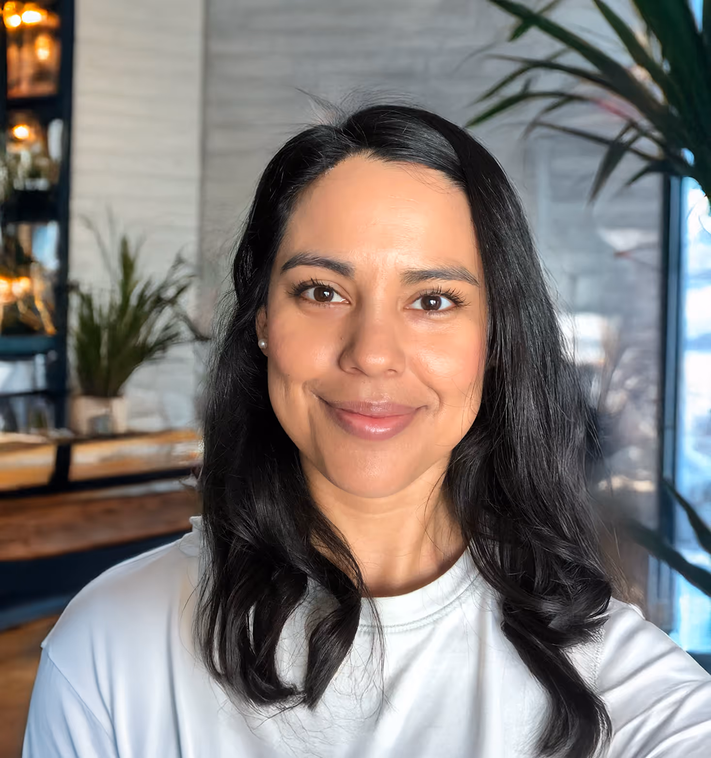 A headshot of a woman with long, dark, wavy hair, smiling softly at the camera. She is wearing a light-colored, possibly white or light gray, t-shirt and small pearl earrings. Her eyes are dark brown. The background is a blurred indoor setting with plants and some shelves visible, suggesting a cafe or a modern office space.