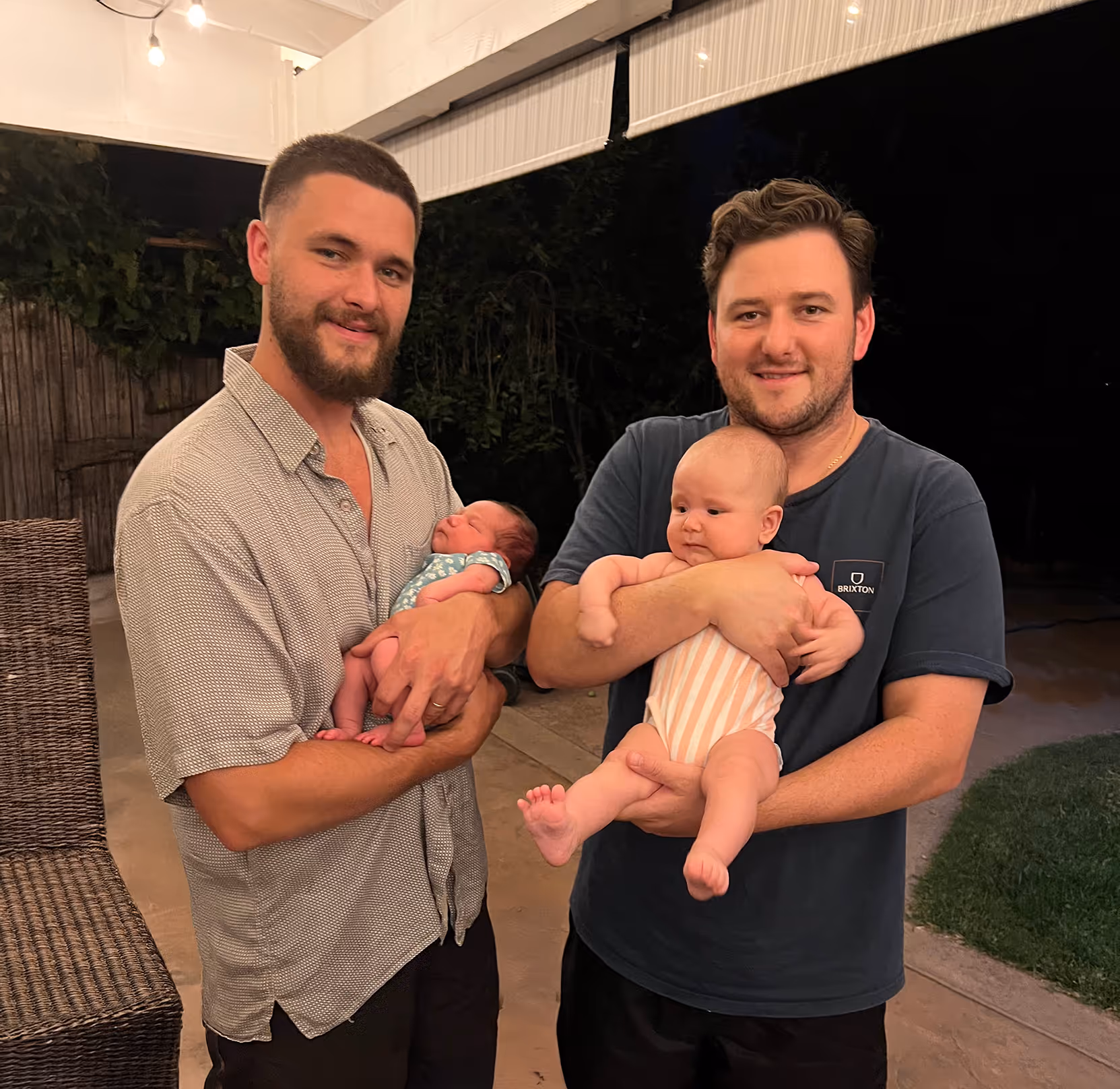 Two men are standing side-by-side outdoors at night, each holding a baby. The man on the right has short hair and a beard, wearing a patterned short-sleeve button-up shirt and dark pants, holding a newborn in his arms. The man on the left has wavy dark hair and a beard, wearing a dark t-shirt, holding a slightly older infant dressed in a striped onesie. Both men are smiling at the camera. In the background, there's a patio area with a wicker chair, a fence, and some greenery. String lights are visible overhead.