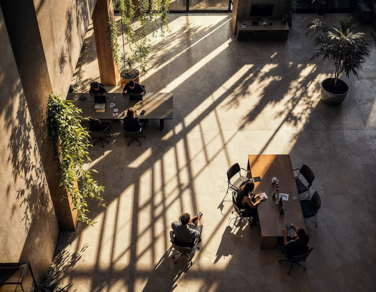 An overhead shot of a spacious, modern interior with high ceilings and large windows, evidenced by the strong diagonal shafts of sunlight and shadows cast across the concrete floor. Several people are scattered throughout the space, working or interacting at large communal tables. Lush green plants hang from the ceiling and sit in pots on the floor, adding to the biophilic design. The overall aesthetic is minimalist with concrete walls and light-colored wooden accents.