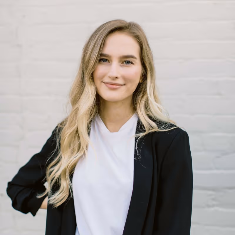 A portrait-style photo of a young woman with long, wavy blonde hair. She is smiling faintly and looking directly at the camera. She is wearing a white t-shirt under a black blazer. The background is a plain white brick wall.