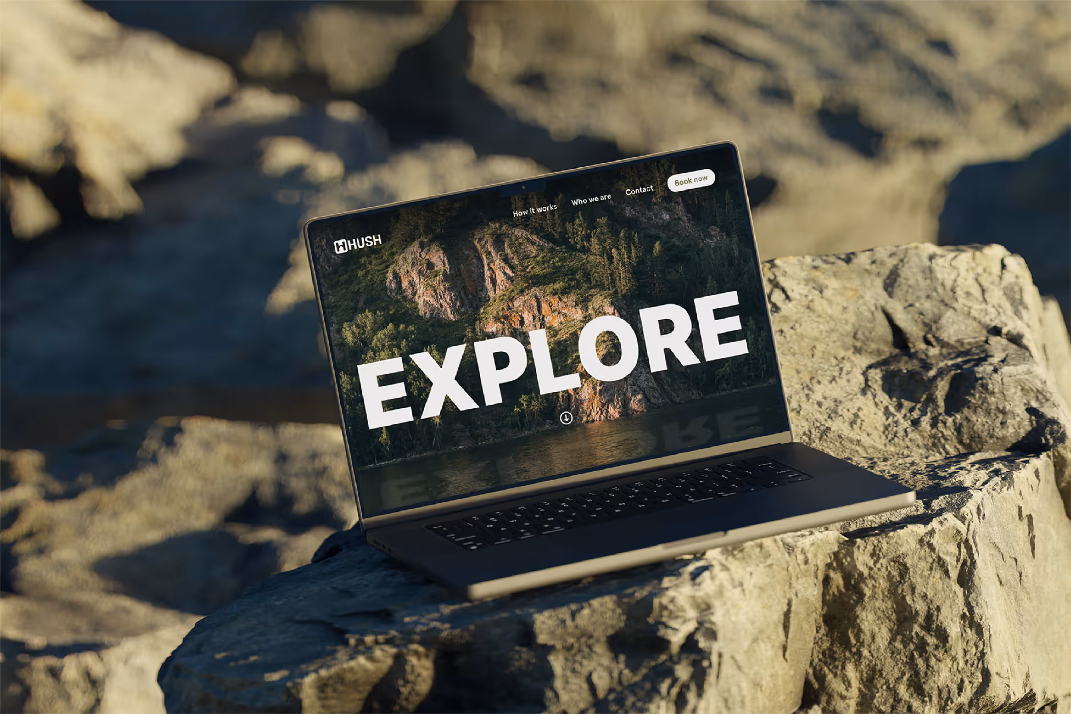 A laptop is open and resting on a rough, rocky surface outdoors, suggesting a natural or rugged setting. The screen displays a website with a large, white text overlay that reads "EXPLORE" against a background image of a mountainous, forested landscape reflected in water. The website also shows navigation links at the top right, including "How it works," "Who we are," "Contact," and a "Book now" button. A logo, "HUSH," is visible in the top left corner. The lighting indicates daylight, with shadows and highlights on the rocks.