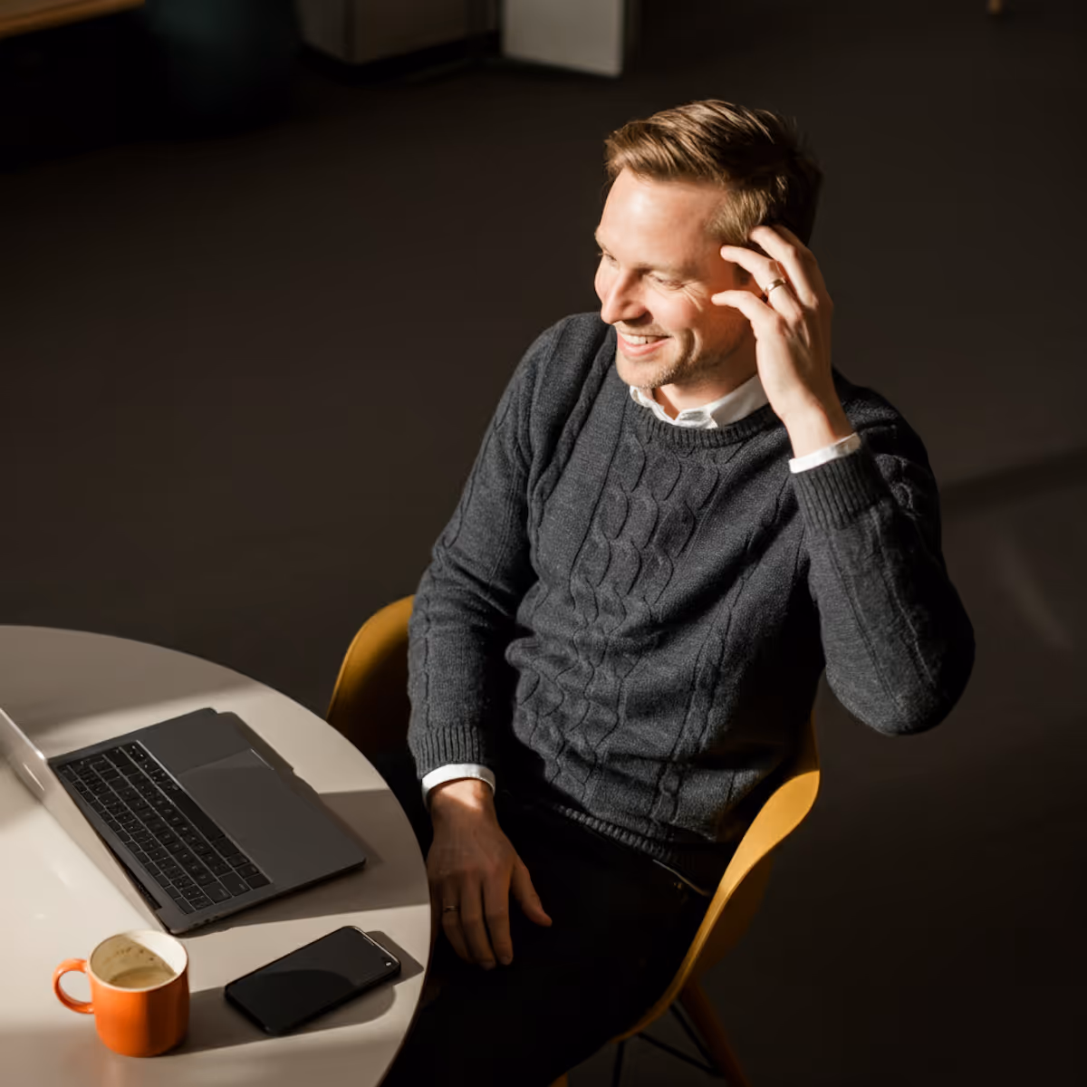 A man with light brown hair is seated at a round white table, smiling and looking off to his left. He is wearing a dark grey cable-knit sweater over a white collared shirt. His left hand is raised to touch his head. On the table, there's a dark laptop, an orange mug, and a black smartphone. He is sitting on a yellow chair. The background is dark and blurred, with light casting a strong, possibly natural, highlight on him and the table.