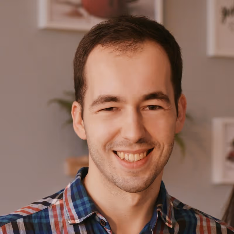 A headshot of a smiling man with short brown hair, looking directly at the camera. He has a slight stubble and is wearing a blue, red, and white plaid shirt. The background is a blurred indoor setting with hints of framed pictures on a wall.