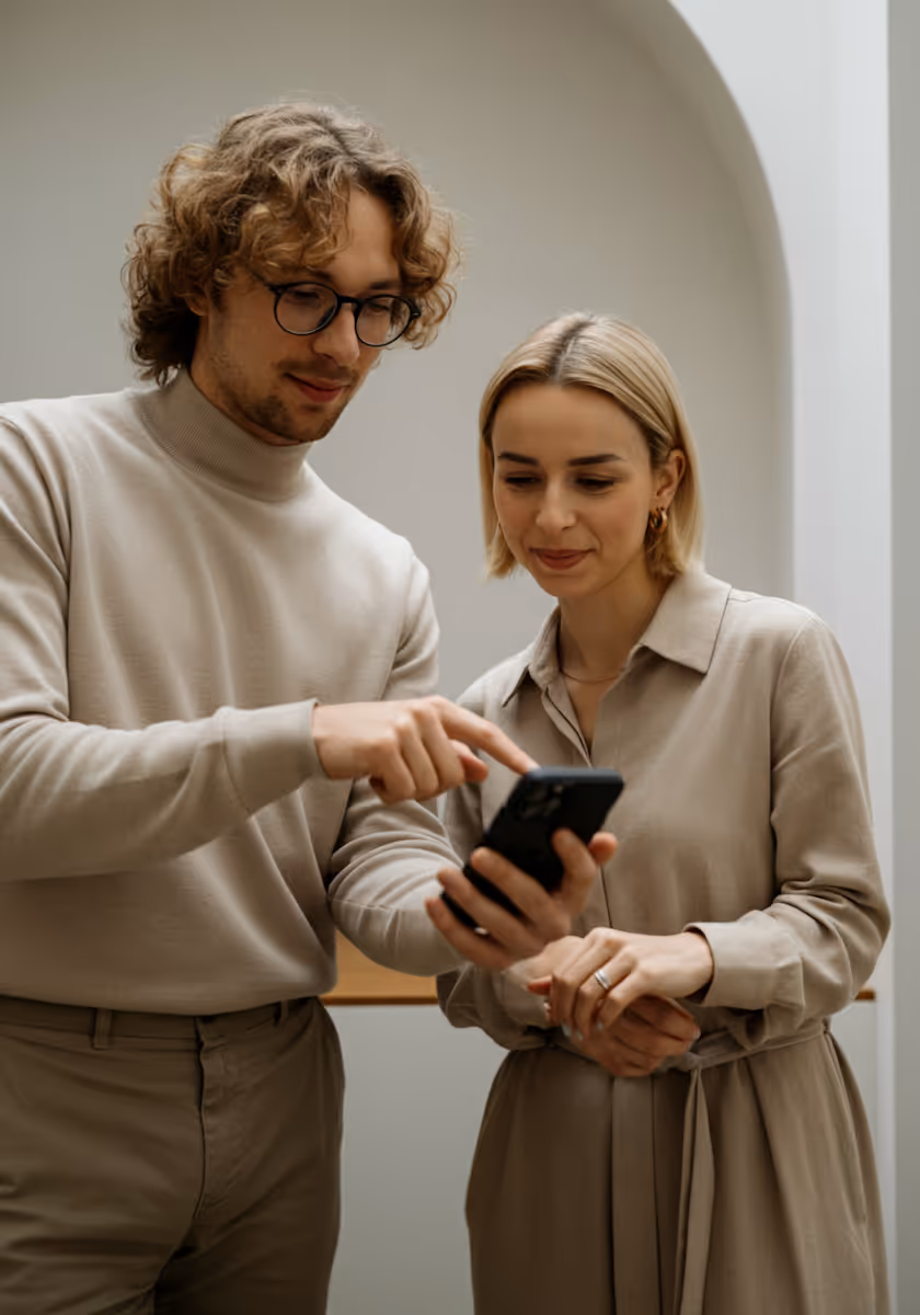 A man and a woman are looking at a smartphone together. The man, with curly light brown hair and glasses, is wearing a beige turtleneck and matching pants, and is pointing at the phone screen. The woman, with short blonde hair and gold hoop earrings, is wearing a beige long-sleeved dress with a tie-waist. They appear to be in an indoor setting with light-colored walls, and an arched doorway is visible in the background.