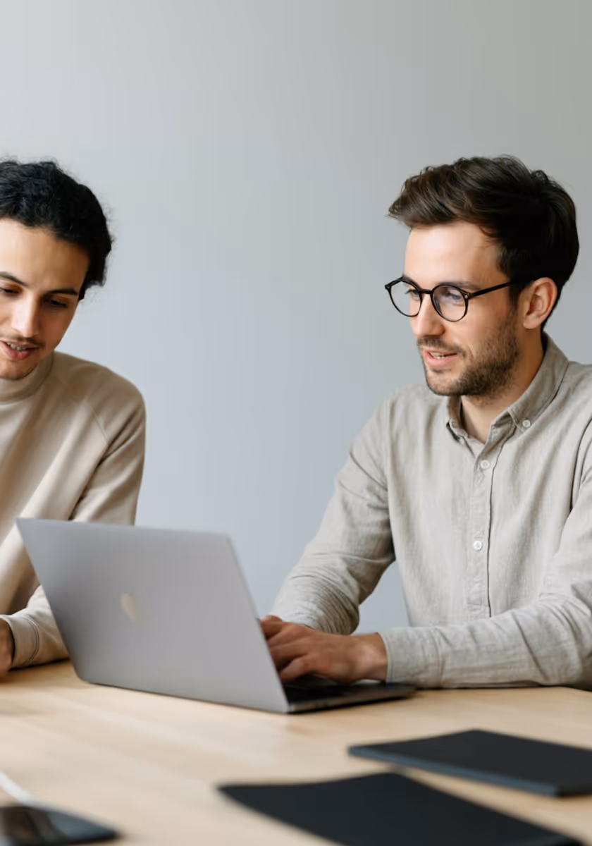 Two men are seated at a light-colored wooden table, looking at a silver laptop. The man on the right, wearing glasses and a light-colored button-up shirt, is actively typing on the laptop. The man on the left, with dark curly hair and wearing a beige turtleneck, is looking towards the laptop screen. The background is a plain light gray wall. There are some dark-colored notebooks or folders on the table in the foreground.