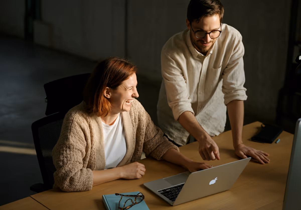 A candid shot of two people, a woman and a man, interacting with a laptop on a wooden desk. The woman, with reddish-brown hair and wearing a light-colored knit cardigan over a white top, is seated at the desk and laughing while looking at the man. The man, with glasses and a light-colored collared shirt, is leaning over the desk, pointing at the laptop screen and smiling. The lighting is dim, with a distinct beam of light illuminating part of the desk and the laptop, creating a warm, focused atmosphere. A pair of glasses and a notebook are also on the desk.
