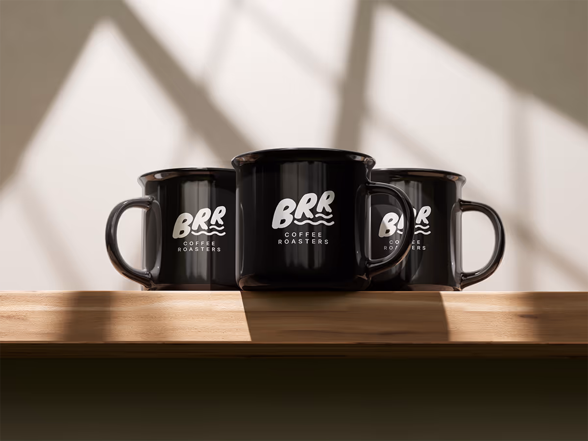 Three black enamel mugs are lined up on a light wooden shelf. Each mug has a white logo that reads "BRR COFFEE ROASTERS." The mug on the far right is slightly out of focus and partially obscured. Sunlight or a strong light source from the left casts distinct shadows of the mugs and diagonal lines from a window or blind onto the white wall behind them.