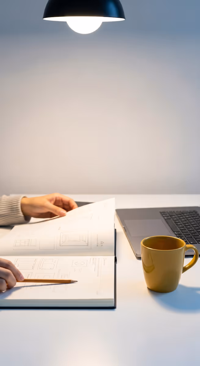 A top-down, cropped shot of a person's hands working at a white desk under a black pendant light. One hand holds open a large notebook or textbook displaying technical drawings or blueprints, while the other holds a pencil, seemingly poised to write or draw. To the right, a dark-colored laptop is partially visible, and a mustard yellow mug sits on the desk. The background is a plain white wall.