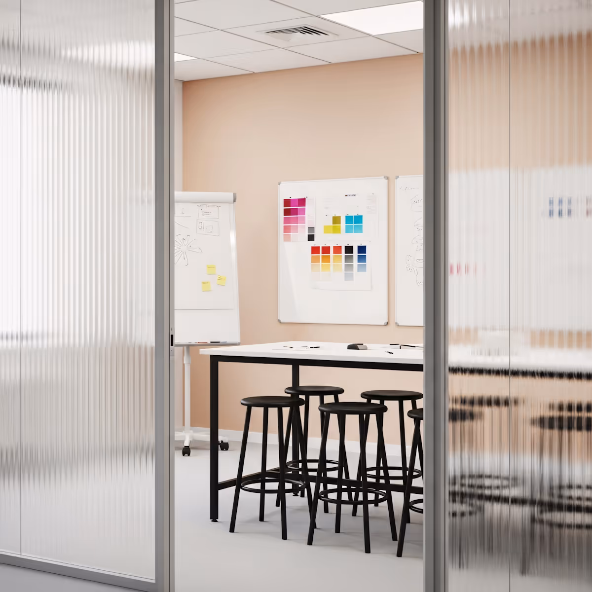 A view into a modern office or meeting room through a fluted glass partition. Inside, there's a light-colored wall, possibly peach or light orange. A long white table is centered, surrounded by several black high-stools. On the wall, there are two whiteboards; one has colorful squares, likely color swatches, and the other has some notes or drawings. A flip chart on a stand is also visible on the left. The ceiling has acoustic tiles and recessed lighting.